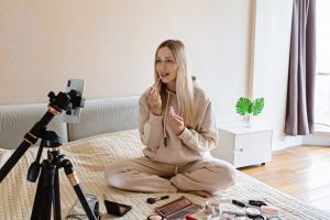 Young Caucasian beauty blogger applying makeup with decorative cosmetics on bed